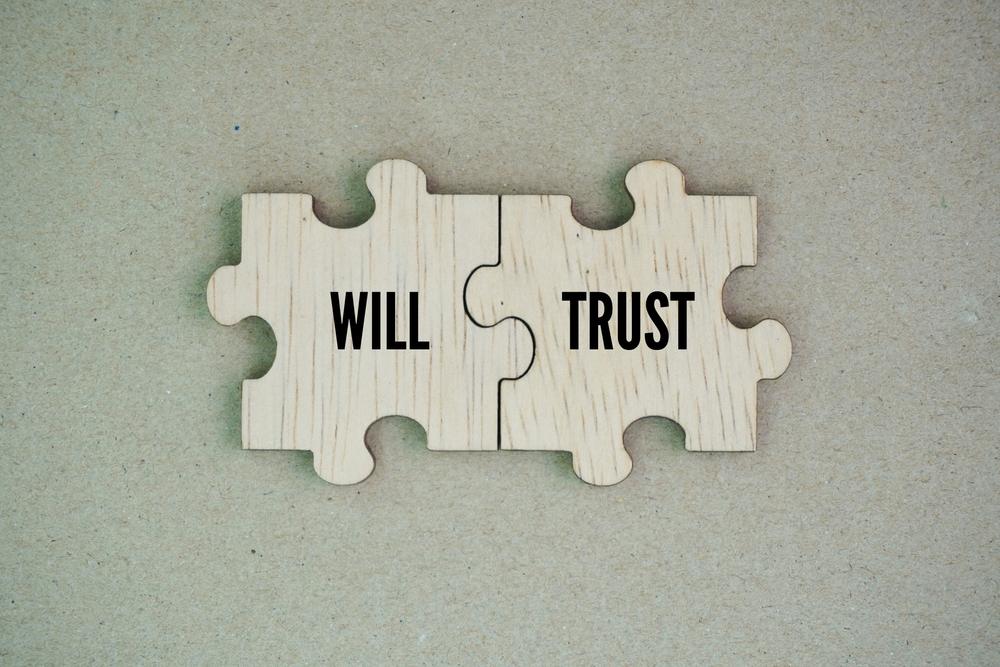 Two wooden puzzle pieces, one of which has “Will” written on it, and another with “Trust” written on it, with someone nearby wondering what the average cost of a will and trust is